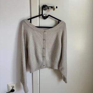 Beige croppad kofta med knappar - Superfin beige kofta med croppad passform och bred ribbad kant. Tröjan har långa ärmar, snygg offshoulder-ringning och knappar framtill. Perfekt att styla med jeans eller kjol för en chill och trendig look.