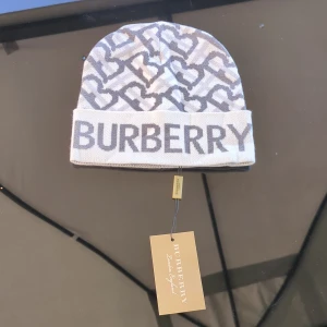 Burberry mössa - Snygg mössa i vit och grå färg med ikoniskt mönster och stor logga framtill. Mjuk och skön, perfekt hela året runt. Klassisk design med uppvikt kant och diskret mönster över hela mössan.