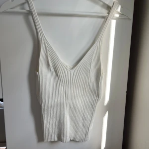 Vit ribbad linnetopp med v-ringning - Säljer en stilren vit ribbad linnetopp med djup v-ringning och smala axelband. Toppen har en figurnära passform och är perfekt för varma dagar. Materialet är mjukt och stretchigt, vilket gör den både bekväm och snygg.