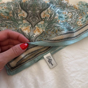 Blommig sidenhalsduk från Aldrich Ave - Tunni siden från Aldrich Ave med blommigt mönster i mintgrönt, beige och inslag av gult. Fyrkantig form med dekorativ kant och detaljerade ornament. Perfekt att knyta runt halsen eller håret för en cool touch.