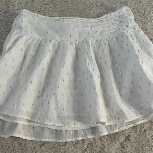 Vit kortkjol med guldprickar Gina Tricot - Supersöt vit kortkjol från Gina Tricot med små glittriga guldprickar över hela tyget. Kjolen har ett brett, veckat midjeband och är lätt utställd med volangig känsla. Perfekt för sommarens alla tillfällen och enkel att matcha med en crop top eller t-shirt.