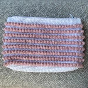 Rosa och vit virkad necessär  - Superfin hammagjord virkad necessär med rosa och vita ränder. Necessären har en mjuk struktur med bubbliga detaljer och stängs med vit dragkedja. Perfekt för att bära småsaker eller smink. Höjd: 12cm, bredd: 20. Du står för frakten, vid mer info skriv 