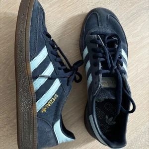 Adidas Spezial sneakers i mörkblå mocka - Adidas Spezial sneakers i mörkblå mocka med ljusblå klassiska tre ränder på sidorna och gummisula. Guldtexten 'Spezial' på sidan ger en retrovibe. Skorna har snörning och rund tå, perfekt för dig som gillar streetstyle.