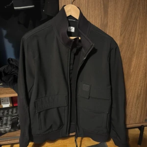 Svart softshelljacka från C.P. Company - Svart softshelljacka från C.P. Company Metropolis Series med C.P. Shell-R-tyg. Jackan har hög krage, stora fickor framtill och diskret logotyp på bröstet. Köpt på NK Göteborg (kvitto finns) skriv för mer bilder 