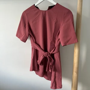 Rosa blus med knyt från Zara Woman - Supersnygg rosa blus från Zara Woman med korta ärmar och knyt i midjan som ger en fin siluett. Blusen har en liten v-ringning framtill och är lätt utsvängd nertill. 