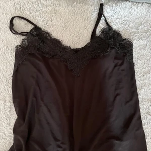 Svart linne från Amisu XS - Supersnygg svart linne från Amisu i satinliknande material med tunna axelband och vacker spetsdetalj upptill. Perfekt för dig som gillar en elegant och stilren look med en touch av romantik.