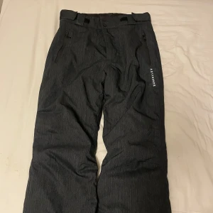 Svarta täckbyxor Sunvalley Typhoon - Snygga svarta täckbyxor från Sunvalley Typhoon-serien med raka ben, dragkedjeförsedda fickor och breda hällor. Nyskick, storlek 38.