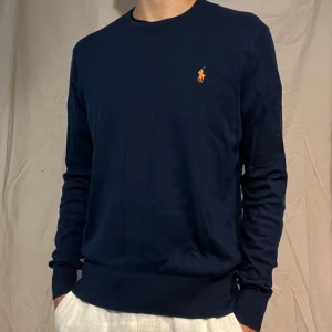 Mörkblå tröja från Polo Ralph Lauren - Mörkblå långärmad tröja från Polo Ralph Lauren, bomull och kashmir med klassisk orange broderad logga på bröstet. Tröjan är i en mjuk mix av bomull och kashmir, har rund hals och ribbade muddar. Perfekt för dig som gillar stilren och tidlös design. Lappen har gått av därav priset, men det syns inte vid användning😊
