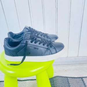 Svarta sneakers från Björn Borg - Svarta sneakers från Björn Borg med klassisk låg profil och vit sula. Skorna har snörning och är tillverkade i skinn med diskret logga på sidan. Perfekta för en clean och stilren look.