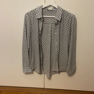 Mönstrad skjorta från ONLY Life, strl 38 - Snygg vit skjorta från ONLY Life i storlek 38 med svart grafiskt mönster. Skjortan har klassisk krage, långa ärmar och knappar framtill. Tunt och lätt material som känns skönt mot huden. Perfekt för dig som gillar stilrena men ändå unika plagg.