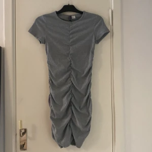 Grå tight kortklänning med rynk - Snygg grå kortklänning med tight passform och rynkade detaljer längs hela framsidan. Klänningen har korta ärmar och rund halsringning, perfekt för dig som gillar en enkel men trendig look.