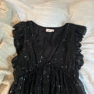Svart glittrig blus från Nelly XS - Supersnygg svart blus från Nelly med v-ringning, volangärmar och massor av paljetter som glittrar. Perfekt för dig som vill sticka ut! Blusen har en figurnära passform.