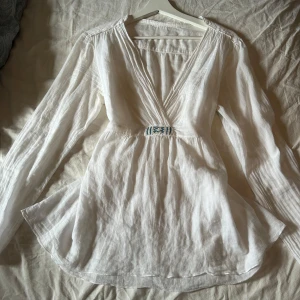 Vit blus med pärldetalj och knyt - Luftig vit blus i bomull med v-ringning och långa ärmar. Snygg pärlbroderad detalj i vitt och blått framtill samt knytband i ryggen för extra passform. Perfekt för en chill och stilren look.