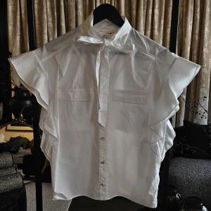 Custommade skjorta - Linora - Snygg vit skjorta i bomull med volangärmar, knyt i halsen och glittriga knappar framtill. Två bröstfickor ger extra stil och skjortan har en luftig, loose passform. Perfekt för dig som vill sticka ut med detaljer och en fräsch look. (Prislapp saknas, men aldrig använd)