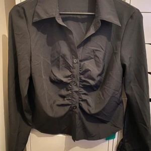 Svart skjorta med rynk från SHEIN - Svart skjorta från SHEIN i storlek M. Skjortan har långa ärmar, klassisk krage och knäppning framtill. Snygg rynkdetalj längs knappraden ger en cool och trendig look. Tillverkad i polyester för en lätt känsla.