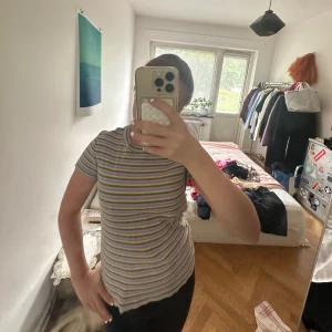 Randig ribbad topp i pastellfärger - Säljer en kortärmad, ribbad topp med smala ränder i lila, grön, gul och vit. T-shirten har en figurnära passform och rund halsringning. Perfekt för dig som gillar färgglada och lekfulla plagg!