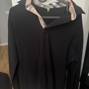 Svart långärmad pikétröja Burberry - Klassisk svart långärmad pikétröja från Burberry med ikoniskt rutigt tyg på insidan av kragen. Tröjan har broderad logga på bröstet och stilren krage. Perfekt för dig som gillar en clean och tidlös look. Passar mest L