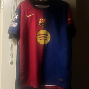 FC Barcelona Raphinha 11 Nike tröja - Säljer en FC Barcelona matchtröja med Raphinha 11 på ryggen. Tröjan är kortärmad och har klassiska blå och röda färger med guldiga detaljer, klubbmärke och sponsorloggor. Materialet är lätt och andas, perfekt för fotboll eller att bära som supporter.