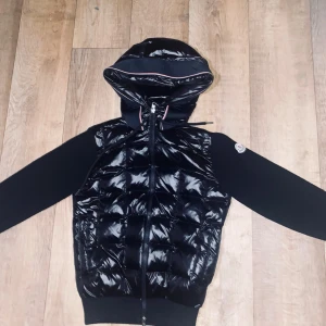 Svart pufferjacka från Moncler - Snygg svart pufferjacka från Moncler med glansig vadderad framsida, stickade ärmar och huva med dragsko. Jackan har dragkedja hela vägen och ikonisk Moncler-logga på ärmen. Perfekt för dig som vill ha en stilren och sportig look.