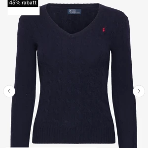  Ralph Lauren tröja - Kabelstickad mörkblå tröja från Polo Ralph Lauren med klassisk v-ringning och röd broderad logga på bröstet. Tröjan har lång ärm och ribbade muddar, perfekt för lager-på-lager och snygg till både jeans och kjol.