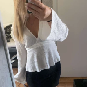 Vit omlottblus med vida ärmar - Supersöt vit omlottblus med v-ringning och långa, vida ärmar. Blusen har knytband i midjan och är gjord i ett lätt, krinklat tyg som ger en chill och luftig känsla. Perfekt att styla med både kjol och jeans för en trendig look. Använda få gånger.💞