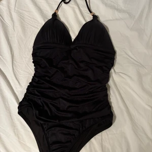 Ny svart baddräkt från Victoria's Secret - Supersnygg svart baddräkt ’the flawless one-piece’ från Victoria's Secret med knyt i nacken och gulddetaljer med VS-logga. Baddräkten har vadderade kupor och lätt rynkade partier framtill för en smickrande passform. Perfekt för stranden eller poolen! Helt ny, aldrig använt. Endast tvättat.