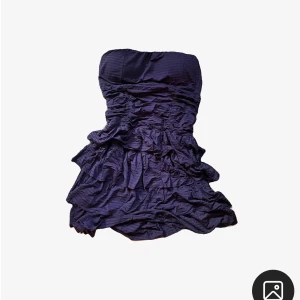 Mörklila axelbandslös kortklänning - Snygg mörklila axelbandslös kortklänning med volangig och rynkad design. Klänningen har en figurnära passform upptill och flera lager i kjolen för extra volym. 