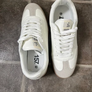 Vita sneakers från 157 med mockadetaljer - Snygga vita sneakers från 157 med beige mockadetaljer på tån och sidorna. Skorna har klassisk snörning, rund tå och en ljusbrun platt sula. Perfekta för dig som gillar stilrena och fräscha sneakers med en retrovibe.