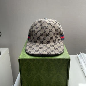 Gucci keps med GG-mönster och läderdetalj - Snygg keps från Gucci i lite blå  med klassiskt GG-monogram över hela kepsen. Har blå och röda ränder på sidorna och justerbar rem i mörkblått läder bak. Tillverkad i bomull och läder, levereras med originalbox och dustbag.