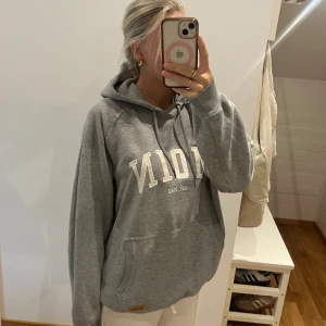 Grå hoodie från Moin med tryck - Säljer en grå hoodie från Moin med vit broderad text framtill och känguruficka. Tröjan har huva med snörning och en avslappnad, loose passform. Perfekt för chill dagar och enkel att matcha med jeans eller mjukisbyxor.