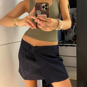 Mörkblå miniskirt - Snygg mörkblå kort omlottkjol med stor ficka på sidan och bälteshällor i midjan. Kjolen har en cool, asymmetrisk design och stängs med knappar på insidan. Perfekt för dig som gillar en stilren men edgy look.