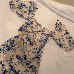 Blommig kortklänning från H&M - Supersöt kortklänning från H&M med beige botten och blått blommönster. Klänningen har puffiga korta ärmar, twistad detalj framtill och cut-out vid midjan. Tillverkad i luftig bomull, perfekt för varma dagar. Romantisk och trendig vibe!