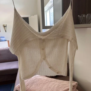 Vit ribbad bolerotopp från Pull&Bear - Superfin vit ribbad bolerotopp från Pull&Bear med knappar framtill och croppad passform. Tunt, stickat material med smala axelband och snygg offshoulder-look. Perfekt att styla över ett linne eller klänning för en trendig vibe.