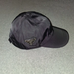 Svart keps från Prada i nylon - Snygg svart keps från Prada i glansig re-nylon med klassisk böjd skärm och ikonisk metallogo på sidan. Kepsen har en stilren look och passar perfekt för dig som gillar exklusiva accessoarer med streetkänsla.