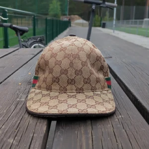Gucci keps med GG-mönster - Snygg keps från Gucci i beige och brunt med klassiskt GG-monogram över hela kepsen. Har gröna och röda ränder på sidorna och justerbar rem baktill i skinn. Perfekt accessoar för att lyfta din streetstyle.