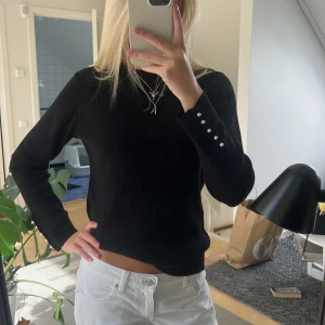 Svart långärmad tröja med pärlknappar - Snygg svart långärmad tröja med pärlknappar på ärmsluten. Enkel och stilren modell som passar perfekt till jeans eller kjol. Tröjan har en rund halsringning och är i ett mjukt material som känns skönt mot huden.