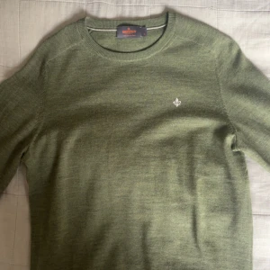 Olivgrön stickad tröja från Morris - Snygg olivgrön stickad tröja från Morris med rund halsringning och diskret logga broderad på bröstet. Tröjan har lång ärm och är tillverkad i mjuk ull, perfekt för lager-på-lager under kyliga dagar.