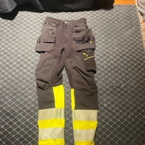 Svarta och gula arbetsbyxor Helly Hansen - Arbetsbyxor från Helly Hansen Workwear i svart och neongult med reflexdetaljer. Flera praktiska fickor, hammarhållare och förstärkta knän. Perfekta för dig som vill synas och ha bra funktion på jobbet. Materialet är slitstarkt och byxorna har smarta detaljer för verktyg. Kniven ingår ej😁 använt en gång bara!