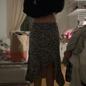 Svart blommig långkjol med volang - Supersnygg svart långkjol med vita småblommor och volangkant. Kjolen har en lätt asymmetrisk nederdel som ger en flowy känsla och passar perfekt till en croppad topp eller stickad tröja. Perfekt för dig som gillar en romantisk och trendig stil.