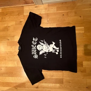 Svart t-shirt från Sweet SKTBS - Svart t-shirt från Sweet SKTBS med stor vit print framtill av en ängel som spelar gitarr och texten 'SWEET SKTBS SKATEBOARDS'. Klassisk passform och rund hals. Perfekt för dig som gillar streetwear och skatekultur.