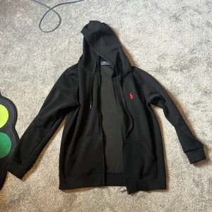 Svart hoodie från Polo Ralph Lauren - Svart hoodie från Polo Ralph Lauren med röd broderad logga på bröstet. Tröjan har huva med snörning, dragkedja framtill och två fickor. Tillverkad i mjuk bomull, perfekt för en chill och stilren look.