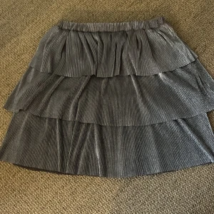 Glittrig silver volangkjol - Supersnygg kortkjol i glittrigt silver med flera volanger och resår i midjan. Kjolen har ett plisserat tyg som ger extra effekt och passar perfekt när du vill sticka ut. Materialet är lätt och skimrande, vilket gör den till ett statement-plagg.