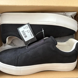 Svarta slip-on sneakers från Exani - Stilrena svarta slip-on sneakers från Exani med vit sula och mjukt mockaliknande material. Skorna har elastisk resår upptill för enkel påtagning och en clean, minimalistisk look. Perfekta för dig som gillar enkel och modern stil. Aldrig använda, ettiketen är kvar.