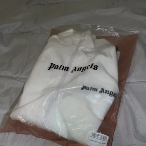 Vit Palm Angels track kofta  - Säljer en clean vit track kofta från Palm Angels med svarta ränder längs ärmarna och logga på bröstet. Kofta har hel dragkedja framtill och ribbade muddar. Perfekt för dig som vill ha en sportig och stilren look.