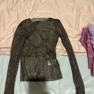 Brun mesh topp med ormmönster - Säljer en transparent brun mesh topp från monki med ormmönster. Tight passform och långa ärmar, perfekt att styla med jeans eller kjol för en edgy look. Tunt och stretchigt material som sitter snyggt på kroppen.