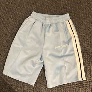 Palm Angels ljusblå shorts med revärer - Palm Angels shorts i ljusblått med vita och svarta revärer längs sidorna. Elastisk midja och sidofickor. Ikonisk Palm Angels-logga tryckt på ena benet. Perfekta för en sportig och trendig look.
