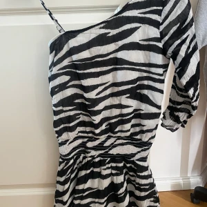 Zebramönstard klänning från Gina Young  - Snygg zebramönstrad kortklänning från Gina Tricot x Izaandelle. Klänningen har en axelbandslös design med en axel och ett smalt axelband, samt trekvartsärm på ena sidan. Den är ganska kort på mig som är 158 cm lång. Den har endast blivit använd 1 gång.