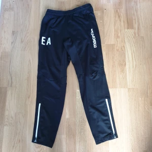 Svarta träningsbyxor Macron XS - Svarta träningsbyxor från Macron i storlek XS. Byxorna har vita detaljer, dragkedjor vid bensluten och snörning i midjan. Macron-logga på benet och tryck med bokstäverna EA. Tillverkade i polyester för en sportig känsla.
