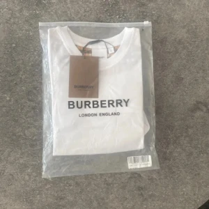 Vit Burberry t-shirt med rutiga ärmar - Vit t-shirt från Burberry med klassiska rutiga korta ärmar i beige, svart och rött. Stor logga framtill med texten 'BURBERRY LONDON ENGLAND'. Skön passform och tillverkad i mjuk bomull. Perfekt för dig som vill ha en clean look med lyxig touch.