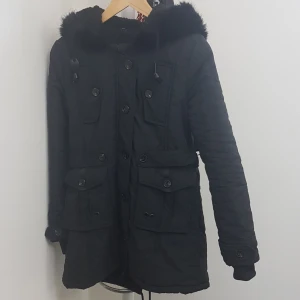 Svart parkajacka med fuskpäls - Svart parkajacka med huva och fuskpäls runt kanten. Jackan har flera stora fickor med knappar, dragkedja och knäppning framtill samt snörning i midjan. Insidan är fodrad med mjuk teddy för extra värme. Perfekt för kalla dagar och snygg till streetwear. PRIS KAN DISKUTERAS!